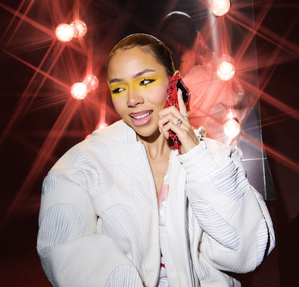 Joven mujer con sombra de ojos amarilla brillante, chaqueta blanca y funda de teléfono roja sonriendo mientras habla por teléfono frente a un fondo con espejo iluminado.