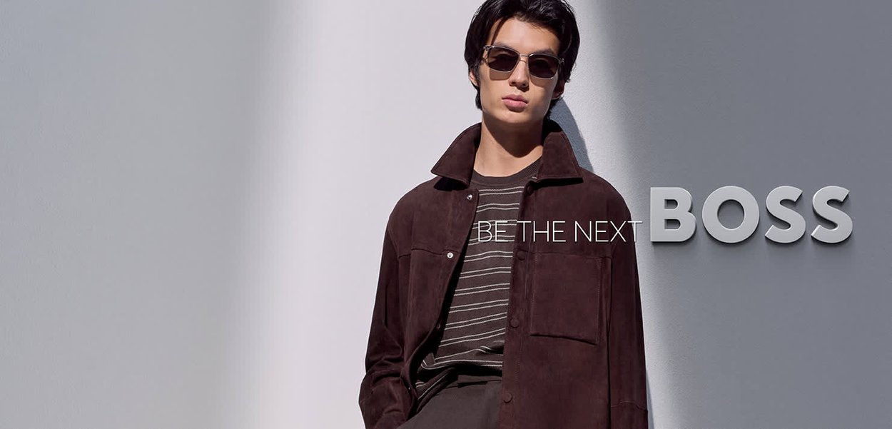 Joven con gafas de sol, chaqueta oscura y camisa a rayas, de pie junto a una pared clara con el texto "SÉ EL PRÓXIMO JEFE" a su lado.