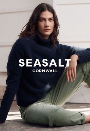 Vrouw met lang donker haar, gekleed in een marineblauwe coltrui en lichtgroene corduroybroek, zittend op de vloer tegen een witte muur.