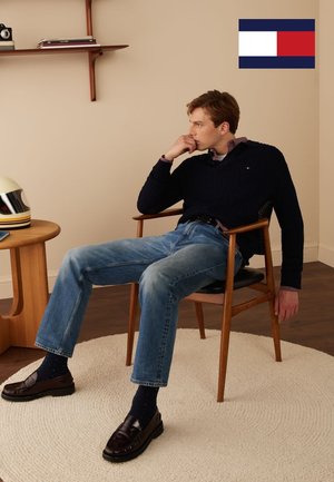 Marineblauwe gebreide trui, geruit overhemd met kraag, lichtblauwe jeans, donkere loafers en marineblauwe sokken met polkadots. Zittend op een houten stoel met een ronde loper.