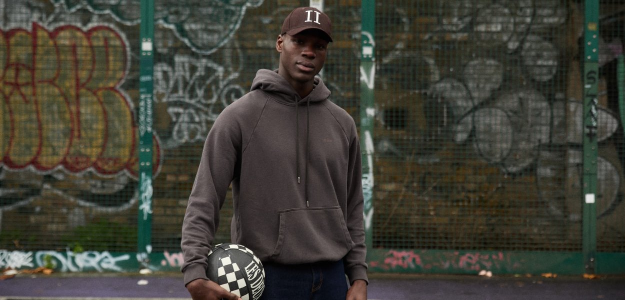 Ung mand i hættetrøje og kasket, der holder en sort-hvidternet fodbold og står foran et hegn med graffiti.