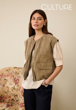 Hellbraune Wildlederweste mit durchgehendem Reißverschluss, kurzen Ärmeln und zwei Vordertaschen. Getragen über einer weißen Bluse, kombiniert mit dunklen Jeans.