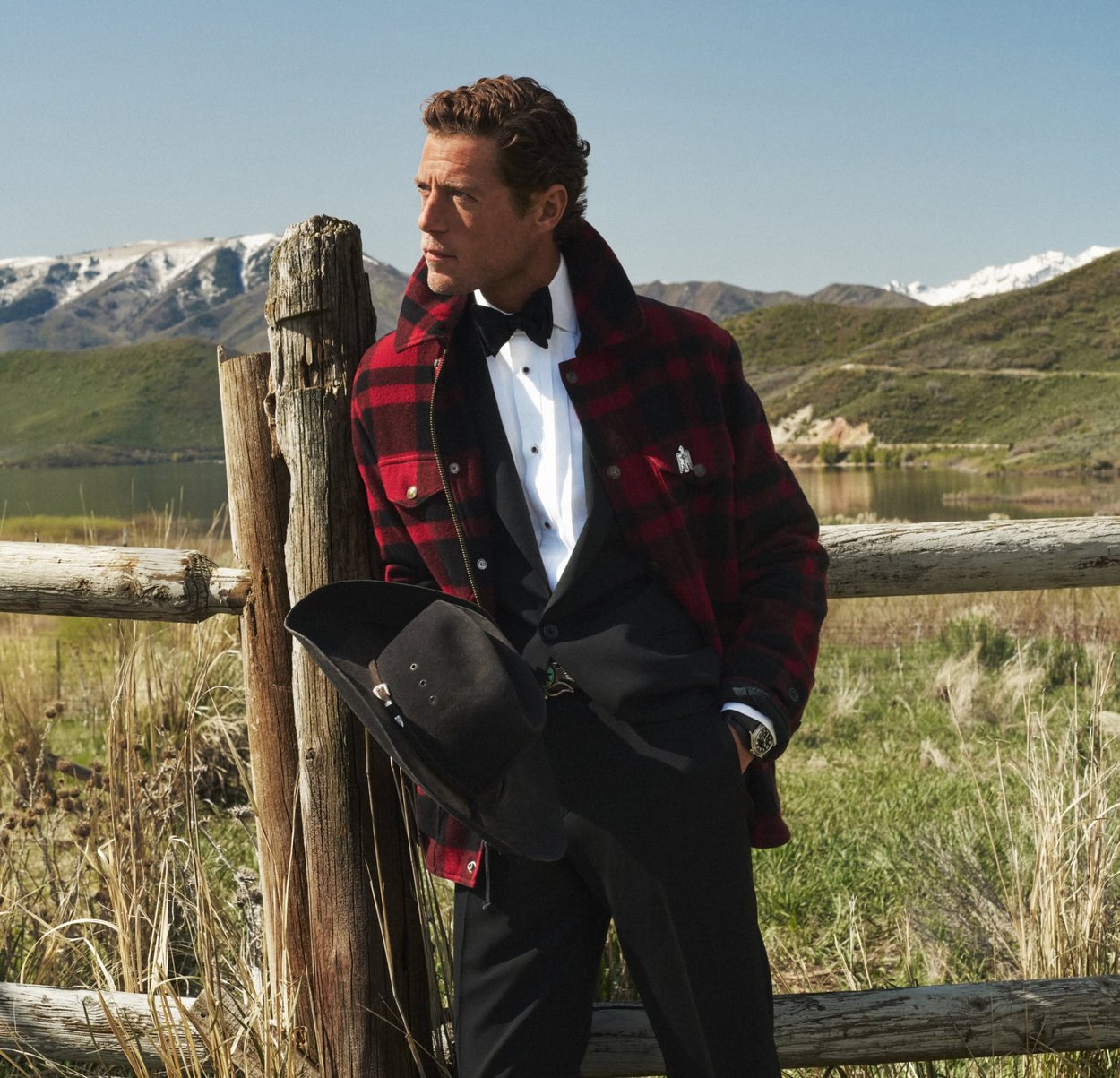 Veste en laine à carreaux rouges et noirs avec un col, superposée à une chemise blanche et un gilet noir. Tenant un chapeau en feutre noir, debout près d'une clôture en bois.