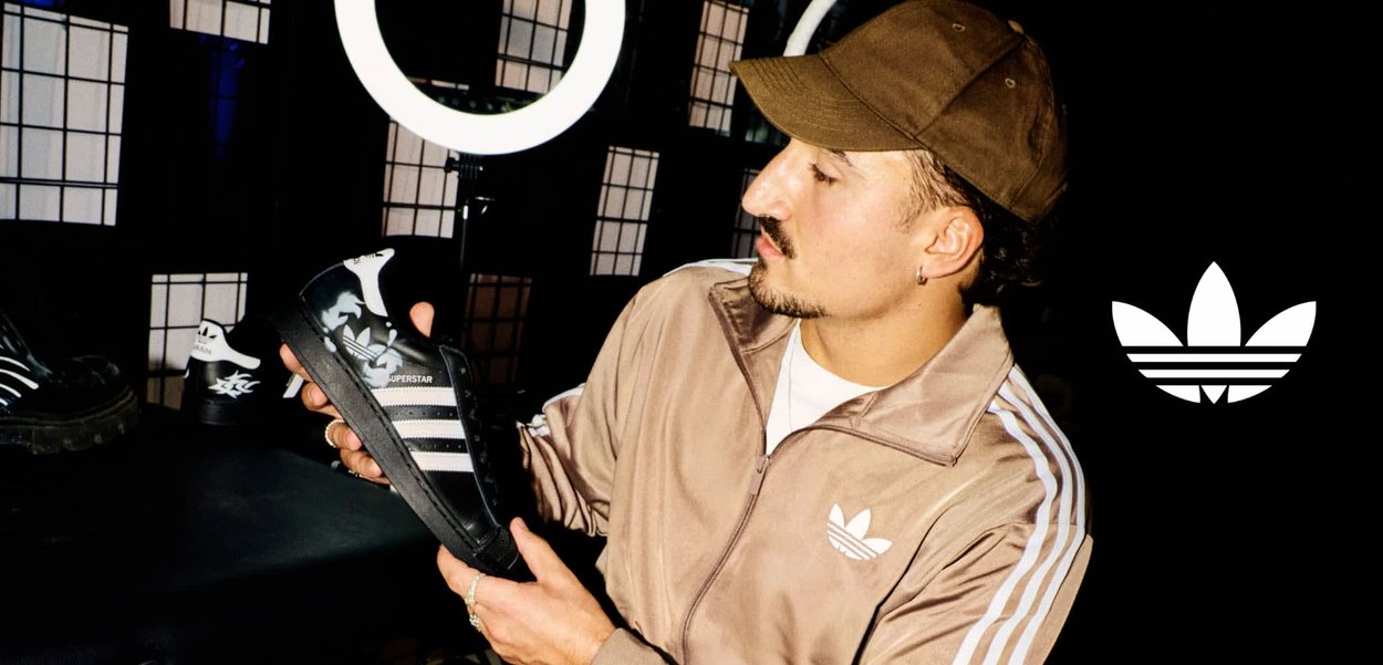 Mann in Adidas-Jacke und -Mütze, der einen schwarzen Adidas Superstar Sneaker in einem Raum mit Schachbrettboden und Ringlicht untersucht.