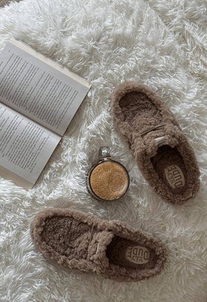 Braune, pelzige Hausschuhe mit strukturierter Oberfläche, gepolstertem Inneren und einem Logoetikett, auf einem weißen flauschigen Teppich neben einer Tasse und einem Buch platziert.