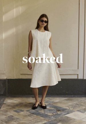 Frau in weißem ärmellosem Kleid, schwarzer Sonnenbrille und schwarzen spitzen Ballerinas steht an einer beigen Wand mit gemustertem Boden.