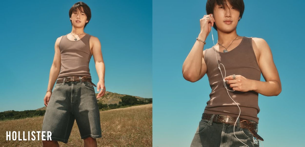 Young man wearing a sleeveless brown top and denim shorts standing outdoors under clear blue sky, holding and adjusting earphones.
