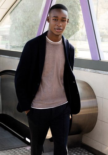 Sort blazer, lys beige sweater og mørke bukser. Glat tekstur med kontrastfarver. Indendørs miljø med moderne arkitektur.