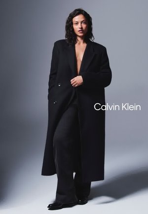 Cappotto in misto lana nero con design lungo e sartoriale, caratterizzato da una chiusura doppiopetto e ampi revers, abbinato a pantaloni neri aderenti.