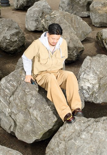 Beige shirt met korte mouwen en zakken, over een grijze hoodie; lichtbruine corduroy broek; zittend op grote grijze rotsen in een buitenomgeving.