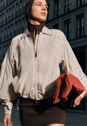 Veste bomber beige clair à coupe décontractée, fermeture éclair frontale et manches tombantes ; le modèle tient un sac rouge, debout devant un bâtiment.