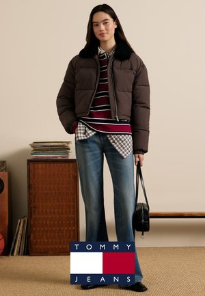 Brązowa kurtka puchowa z czarnym futrzanym kołnierzem, nałożona na pasiastą koszulę w kratkę, do tego jasnoniebieskie dżinsy i czarna torba.