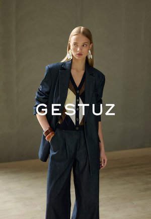 Vrouw met een oversized donkerblazer en wijde broek, gecombineerd met een button-down vest met ruitpatroon en grote witte bladvormige oorbellen.