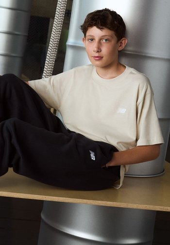 Ein hellbeiges T-Shirt mit einem kleinen weißen Logo, kombiniert mit schwarzen Jogginghosen, die ein weißes Logo aufweisen, sitzt auf einer Holzoberfläche.