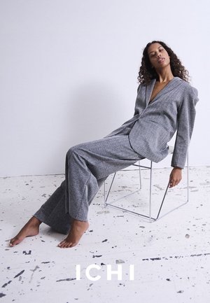 Grau strukturierter Anzug mit einem tief ausgeschnittenen Blazer und Hosen mit weitem Bein. Modell sitzt auf einem Drahtstuhl vor einem hellen Hintergrund.