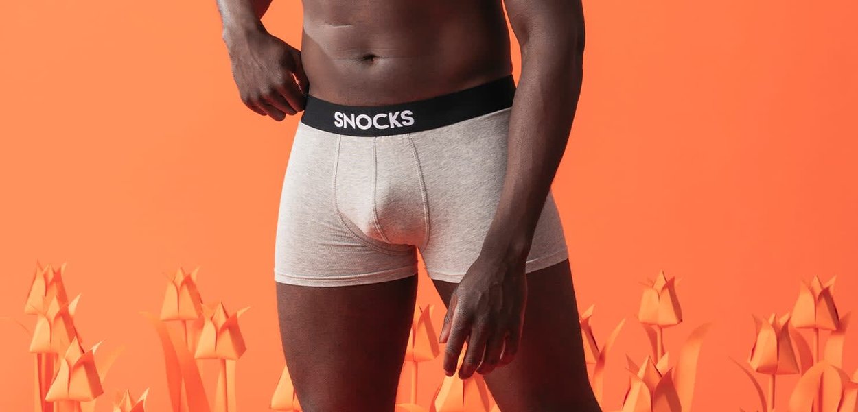 Mandekrop iført lysegrå boxershorts med sort taljebånd mærket "SNOCKS", sat mod en orange baggrund med stiliserede blomster.