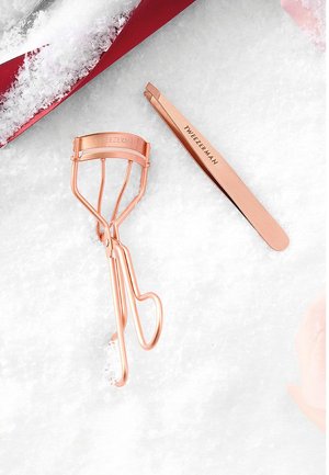 Recourbe-cils en métal et pinces à épiler en rose doré. Le recourbe-cils présente un design ouvert avec une tête incurvée ; les pinces à épiler ont une pointe pointue et un logo gravé.