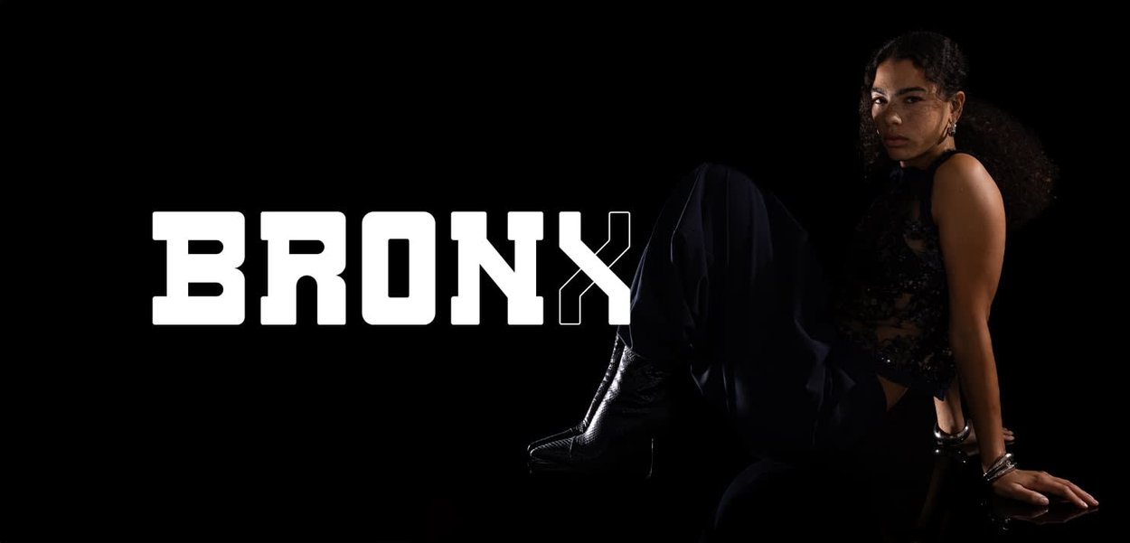 Frau in dunkler Kleidung und Stiefeln sitzt vor schwarzem Hintergrund; links von ihr fettgedruckter weißer Schriftzug "BRONX".