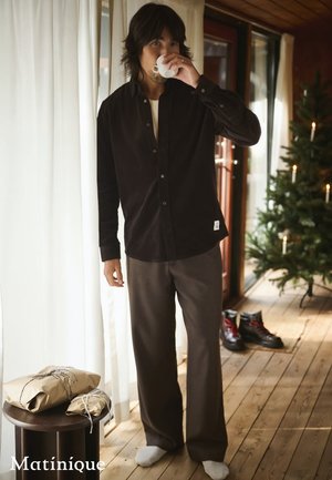 Black corduroy button-up shirt worn with gray pants. Wooden floor, white curtains, and a green Christmas tree in background. Holding a white cup.