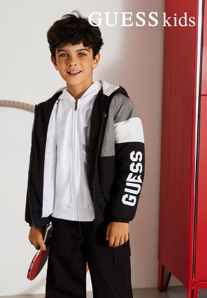 Boy wearing black, white, and gray jacket with "GUESS" on sleeve, holding a table tennis paddle, standing indoors near a red cabinet.