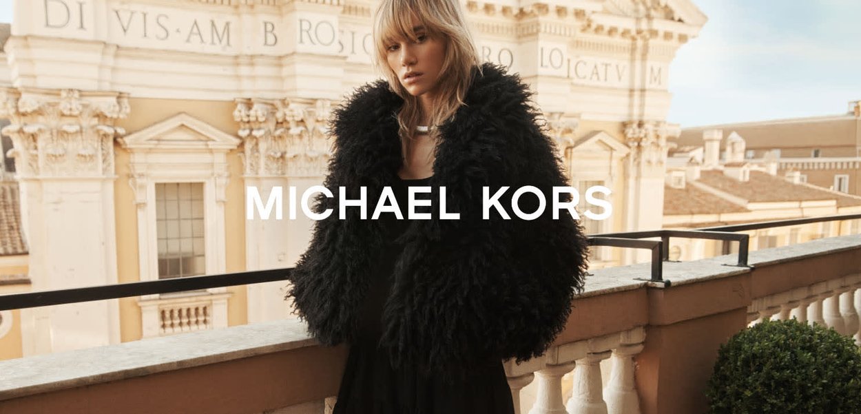 Kvinne iført en svart fluffy kåpe står på en balkong foran en klassisk bygning med arkitektoniske detaljer.