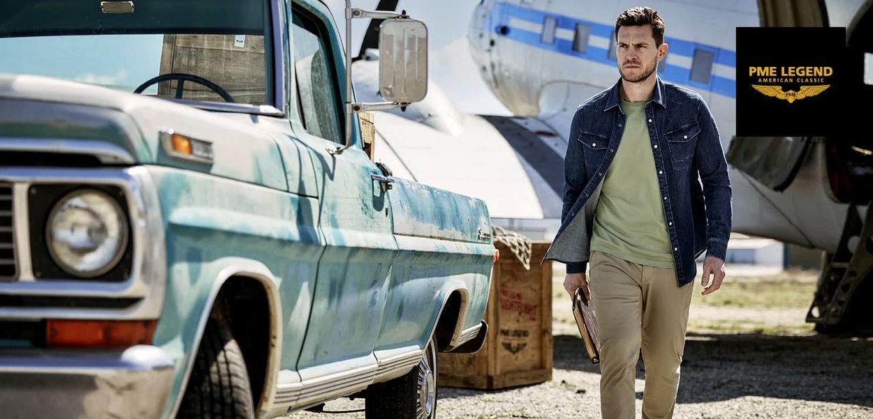 Camionnette bleue vintage avec des accents blancs ; un homme en veste en denim et chemise verte tient une mallette brune, marchant dans une zone ouverte.