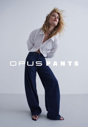 Vrouw met golvend blond haar die een witte blouse en donkerblauwe wijde broek met patroon draagt, staat tegen een effen lichte achtergrond.