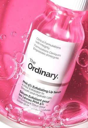 Clear bottle containing pink lip serum, labeled "PHA 5% Exfoliating Lip Serum," with white cap and bubbles in a pink liquid background.