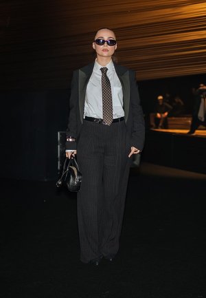 Mujer con blazer negro de gran tamaño, camisa blanca, corbata de lunares, pantalones anchos de rayas, gafas de sol, sosteniendo un bolso negro, de pie en un espacio con poca luz.