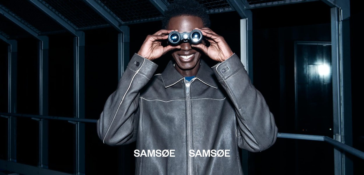 Smilende person i en grå jakke kigger gennem en kikkert om natten på en metalbro eller gangsti med en mørk baggrund.