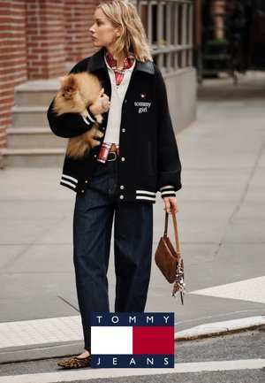 Chaqueta varsity negra con rayas blancas, camisa de cuadros roja, pantalones de mezclilla, llevando un pequeño perro pomerania y un bolso marrón.