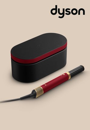 Strumento per lo styling dei capelli Dyson in rosso e oro con manico nero accanto a una custodia nera e rossa su sfondo beige.