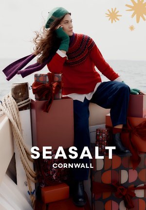 Roter Strickpullover mit gemustertem Passe, dunkelblaue, weit geschnittene Hosen, grüne Handschuhe und passender Hut, umgeben von eingepackten Geschenken.