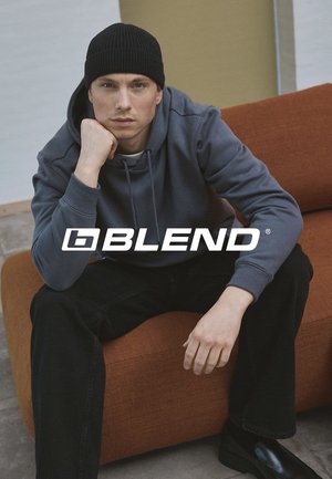 Grå hættetrøje med en ribbet sort beanie. Model siddende på en orange tekstureret sofa med sorte jeans og skinnende sko, poserer eftertænksomt.