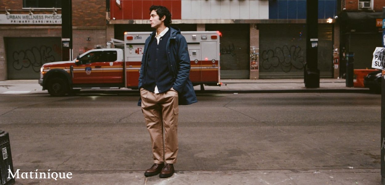 Lässiges Outfit mit einem langen marineblauen Mantel über einem weißen Hemd, beigen Cargohosen und dunklen Schuhen, vor einem urbanen Hintergrund.