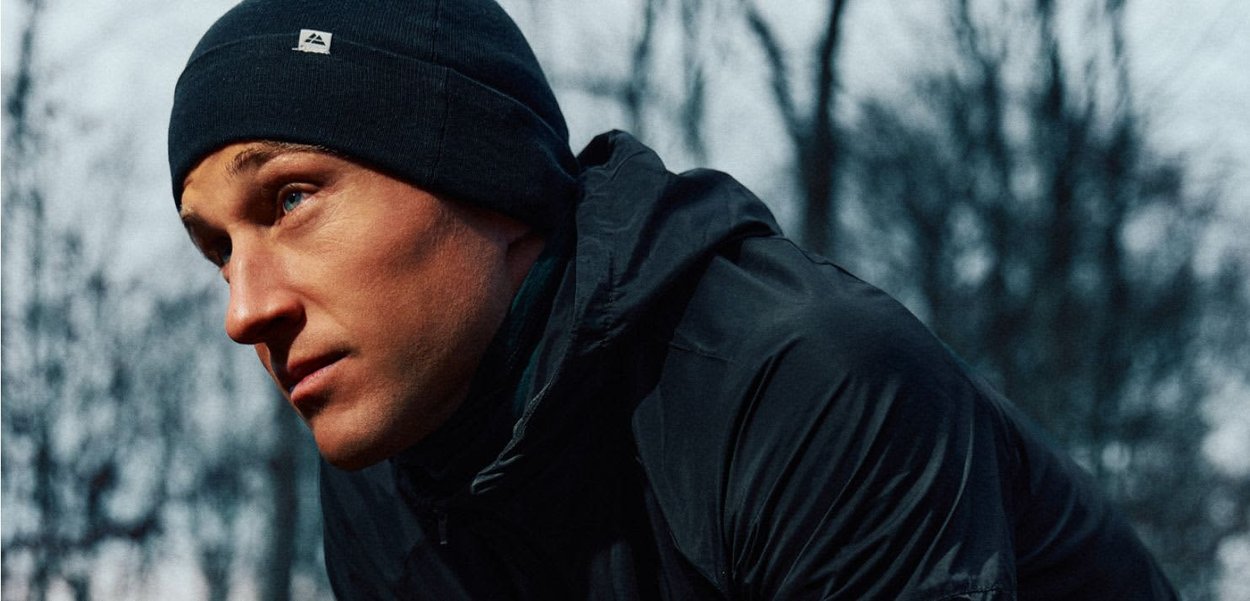 Młody mężczyzna w czarnej czapce i kurtce patrzy uważnie przed siebie na zewnątrz, w tle rozmyte, bezlistne drzewa.