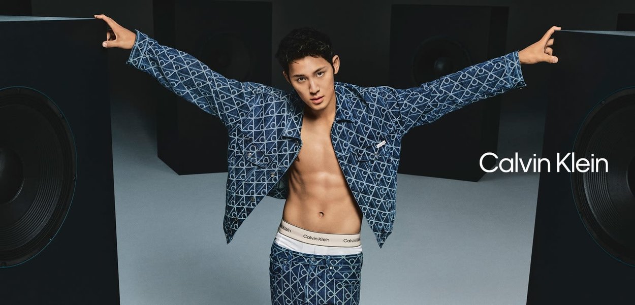 Veste en denim et pantalon avec un motif géométrique bleu, avec une taille marquée Calvin Klein en blanc, posés devant de grands haut-parleurs sombres.