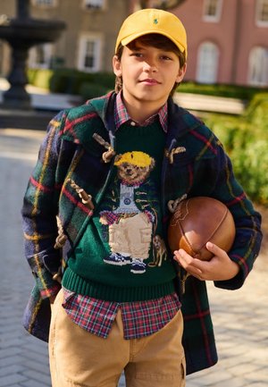 Ein Kind trägt einen dunklen karierten Mantel über einem grünen Pullover mit einem Bärenmotiv, ein kariertes Hemd, khakifarbene Shorts und hält einen braunen Football.