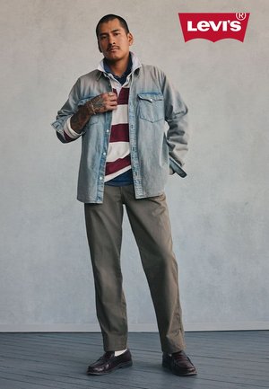 Mann trägt gestreiftes Hemd, hellblaue Jeansjacke, braune Hose und dunkle Schuhe, steht vor einer schlichten Wand, darüber das Levi's-Logo.