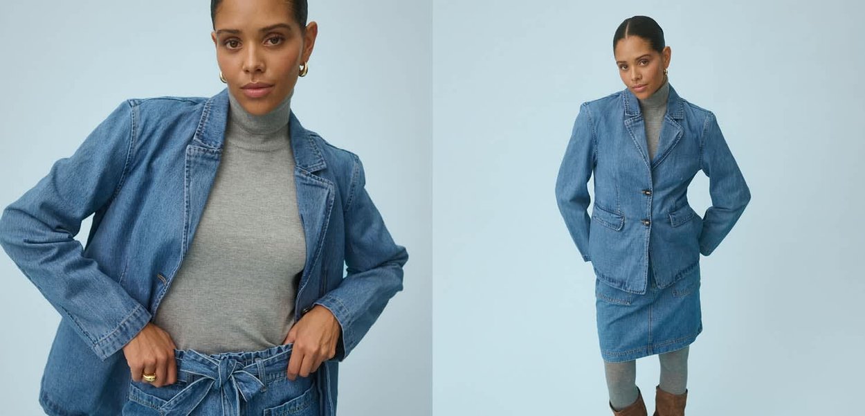 Frau trägt Jeansblazer und Rock über grauem Rollkragenpullover, posiert vor schlichtem hellblauem Hintergrund.