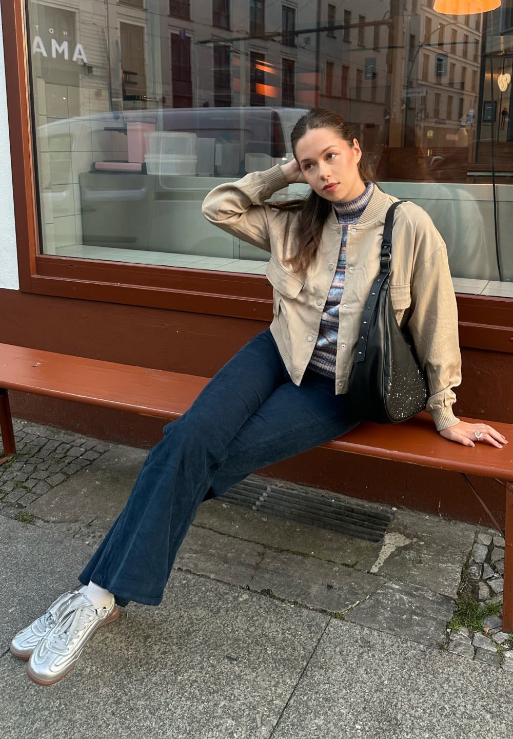 Beige bomber jacket, striped turtleneck, dark blue flared pants, silver sneakers, and a black bag with studs. Sitting on a brown bench.