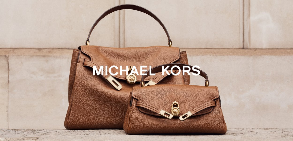 Dos bolsos de cuero marrón de Michael Kors con herrajes en tono dorado están expuestos sobre una superficie de piedra frente a una pared beige.