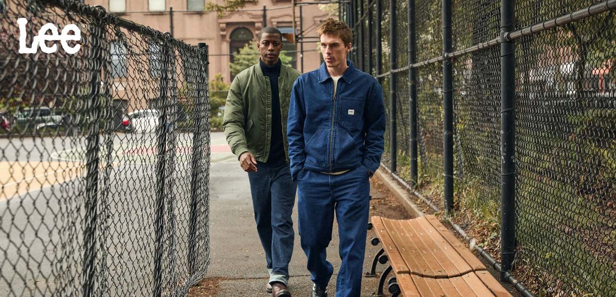 To personer går langs en sti ved en kjede-lenke gjerde. Den ene har på seg en grønn bomberjakke og jeans, den andre en blå denimjakke og matchende bukser.