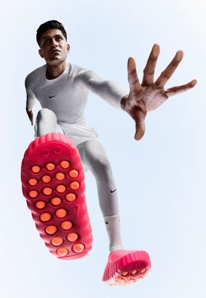Nike Sportschuh mit erhöhter pinkfarbener Sohle und orangen runden Grip-Elementen; Modell in weißer Kleidung streckt sich nach vorne und zeigt dynamische Bewegung.