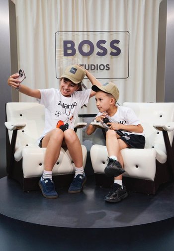 Deux enfants portant des casquettes et des t-shirts blancs sont assis sur des chaises blanches, l'un prenant un selfie et l'autre tenant un microphone chez Boss Studios.