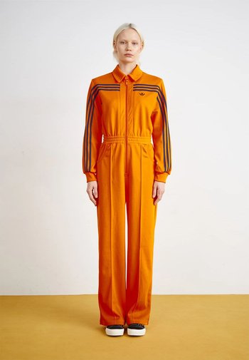 Oranje trainingspak met zwarte strepen, voorzien van een kraag, rits aan de voorkant en elastische tailleband. Losse pasvorm, lange mouwen en wijde pijpen.