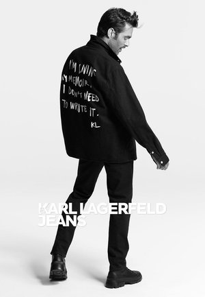 Veste en denim noire avec texte blanc dans le dos, assortie à un jean noir et des bottes noires imposantes. Modèle posé de profil sur un fond clair.