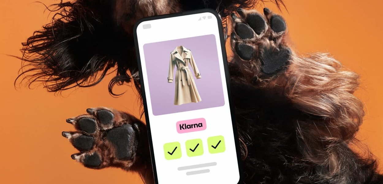 Beige Trenchcoat, präsentiert auf einem Smartphone mit lila Hintergrund. Das Telefon zeigt drei grüne Häkchen und das Logo "Klarna". Ein brauner Hundepfotenabdruck ist sichtbar.
