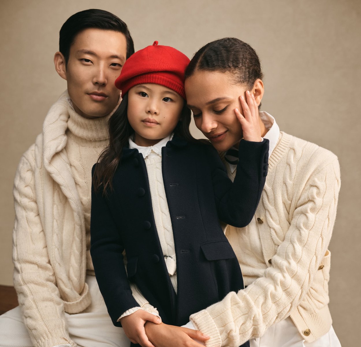 Trois personnes posent ensemble. L'enfant porte un manteau marine et un chapeau rouge, tandis que les adultes portent des pulls en tricot torsadé crème. Fond neutre.