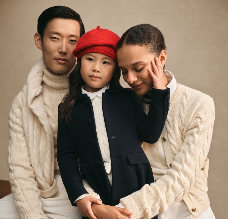Trois personnes posent ensemble. L'enfant porte un manteau marine et un chapeau rouge, tandis que les adultes portent des pulls en tricot torsadé crème. Fond neutre.